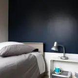 Chambre bleu nuit et taupe : un lit douillet avec un oreiller blanc et gris, une lampe de bureau argentée et une tasse bleue sur une table de chevet blanche.