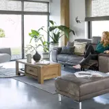 Une femme se détend dans un salon cocooning, avec un fauteuil confortable, un canapé en cuir et beaucoup de plantes vertes.