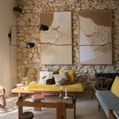 Un salon ancien moderne avec des tapisseries texturées sur un mur de pierre, des meubles en rotin et des coussins jaune vif.