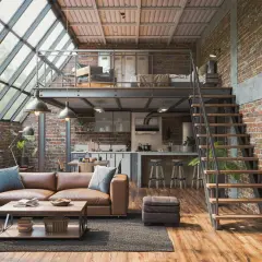 Un grand salon de style loft industriel avec des murs en briques apparentes, un canapé en cuir marron, une mezzanine avec un lit et une cuisine ouverte.