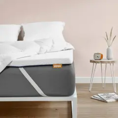 Un matelas trop mou que faire ? Ce surmatelas blanc sur un matelas gris offre une solution. Un réveil orange et des magazines sont sur une table de chevet.
