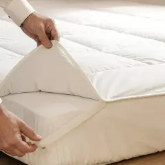 Un homme ajuste un surmatelas blanc sur un matelas. Il sert à améliorer le confort et la protection de votre literie.
