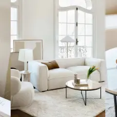 Un salon sans tv, ambiance cosy avec canapé blanc, table basse en marbre et lampes. Des livres et des vases décorent l'espace.