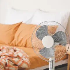 Ventilateur sur pied prêt à rafraîchir une pièce, avec un lit défait en arrière-plan.