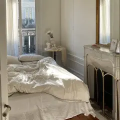 Une petite chambre cocooning baignée de soleil. Le lit défait invite au repos, une cheminée ornée de bibelots et un miroir doré complètent ce tableau paisible.
