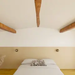 Chambre mansardée aux poutres apparentes, peinte en deux couleurs : blanc et beige. Le lit est fait, avec un coussin décoratif.