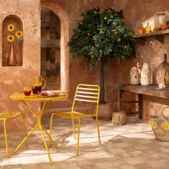 Terrasse ensoleillée aux couleurs terracotta et jaune. Table et chaises jaunes, oranger, tournesols et vaisselle en verre.