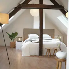 Une chambre minimaliste sous les combles, avec un lit blanc douillet, des poutres apparentes et une plante verte.