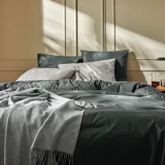 Un lit douillet avec un drap en soie ou satin anthracite, des coussins gris et une couverture douce. La lumière du soleil crée des ombres sur le mur.