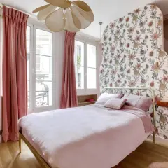 Chambre rose poudré et bois, avec un lit douillet, des rideaux velours, un papier peint floral et un lustre original.