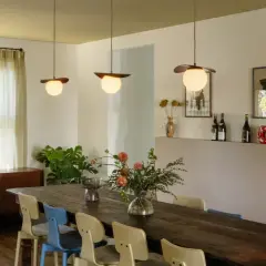 Trois luminaires au-dessus d'une grande table en bois, éclairant un bouquet de fleurs et des chaises colorées.