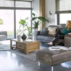 Une femme se détend dans un salon cocooning, avec un fauteuil confortable, un canapé en cuir et beaucoup de plantes vertes.