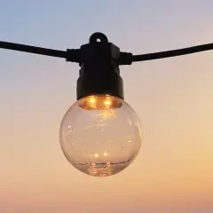 Une ampoule ronde, éclairée, suspendue à un fil noir, se reflète dans le ciel doux du soir. IP 45.
