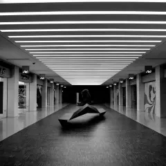 Un couloir de centre commercial, éclairci par des néons, avec une sculpture moderne au centre. Les boutiques sur les côtés sont sombres, créant un contraste saisissant.