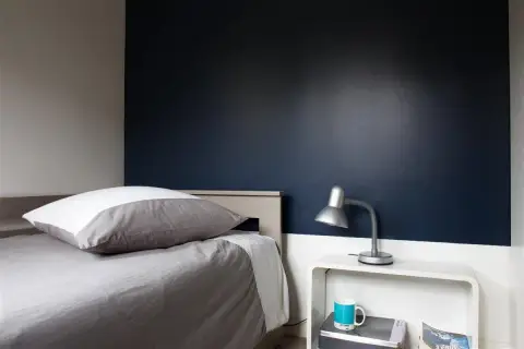 Chambre bleu nuit et taupe : un lit douillet avec un oreiller blanc et gris, une lampe de bureau argentée et une tasse bleue sur une table de chevet blanche.