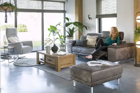 Une femme se détend dans un salon cocooning, avec un fauteuil confortable, un canapé en cuir et beaucoup de plantes vertes.