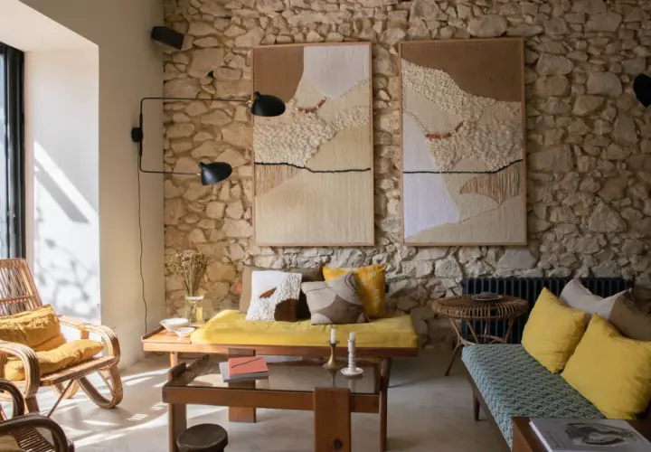 Un salon ancien moderne avec des tapisseries texturées sur un mur de pierre, des meubles en rotin et des coussins jaune vif.