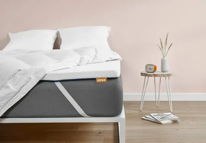Un matelas trop mou que faire ? Ce surmatelas blanc sur un matelas gris offre une solution. Un réveil orange et des magazines sont sur une table de chevet.