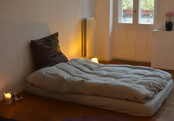 Un matelas au sol sans sommier, prêt pour une nuit paisible. Une bougie allumée éclaire doucement la pièce.