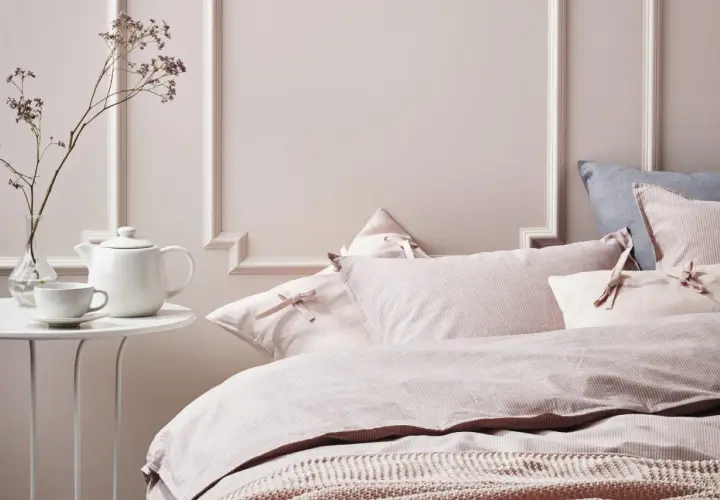 Chambre couleur pastel mur avec un lit douillet, des coussins roses à rayures et une théière blanche sur une table.