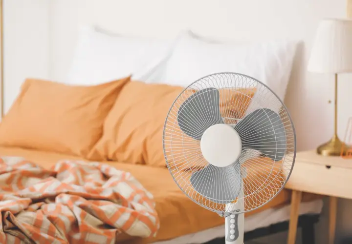 Ventilateur sur pied prêt à rafraîchir une pièce, avec un lit défait en arrière-plan.