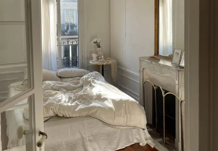 Une petite chambre cocooning baignée de soleil. Le lit défait invite au repos, une cheminée ornée de bibelots et un miroir doré complètent ce tableau paisible.