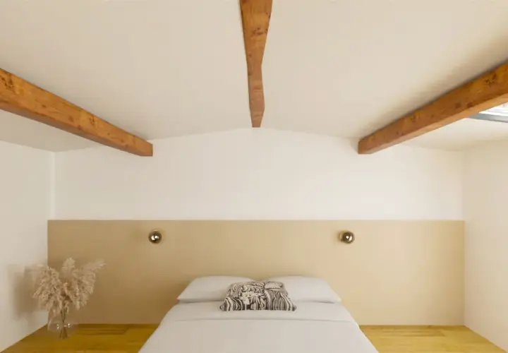 Chambre mansardée aux poutres apparentes, peinte en deux couleurs : blanc et beige. Le lit est fait, avec un coussin décoratif.