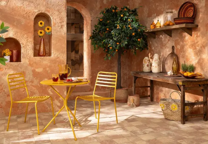 Terrasse ensoleillée aux couleurs terracotta et jaune. Table et chaises jaunes, oranger, tournesols et vaisselle en verre.