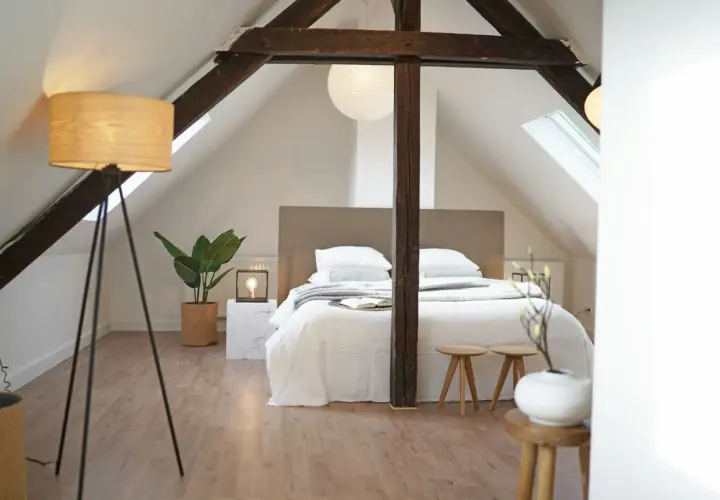 Une chambre minimaliste sous les combles, avec un lit blanc douillet, des poutres apparentes et une plante verte.