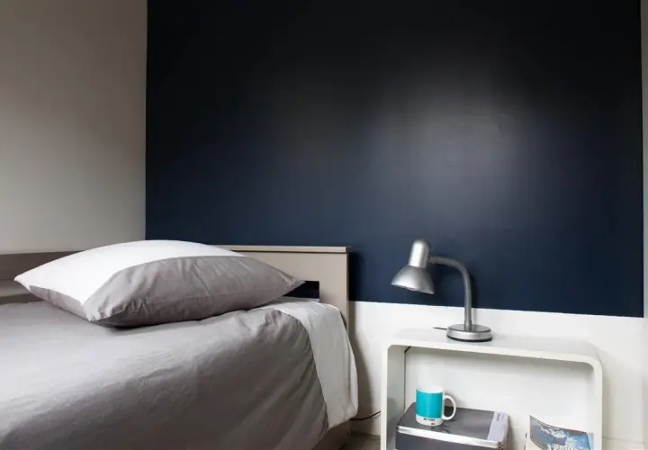 Chambre bleu nuit et taupe : un lit douillet avec un oreiller blanc et gris, une lampe de bureau argentée et une tasse bleue sur une table de chevet blanche.