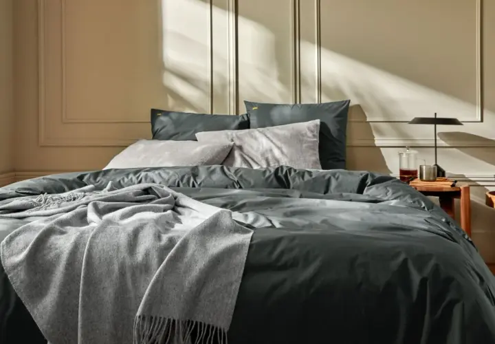 Un lit douillet avec un drap en soie ou satin anthracite, des coussins gris et une couverture douce. La lumière du soleil crée des ombres sur le mur.