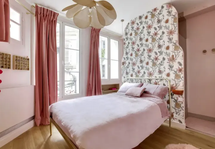 Chambre rose poudré et bois, avec un lit douillet, des rideaux velours, un papier peint floral et un lustre original.