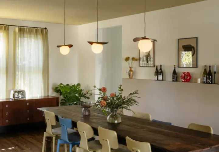 Trois luminaires au-dessus d'une grande table en bois, éclairant un bouquet de fleurs et des chaises colorées.