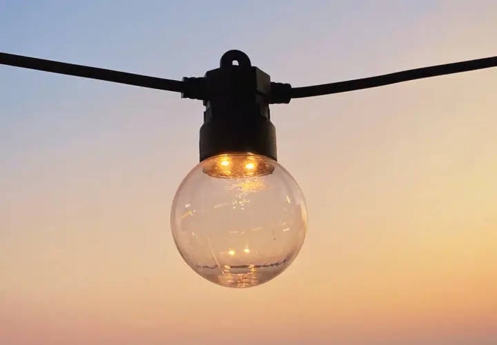Une ampoule ronde, éclairée, suspendue à un fil noir, se reflète dans le ciel doux du soir. IP 45.