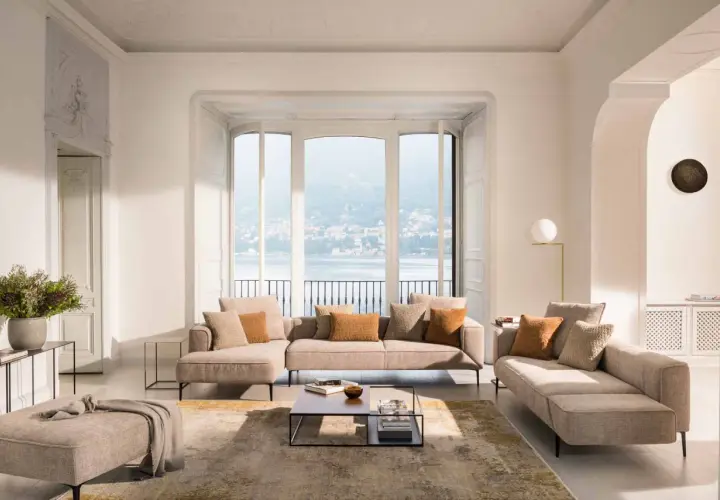 Un salon cocooning moderne avec canapé modulable, pouf, tapis et vue sur un lac.