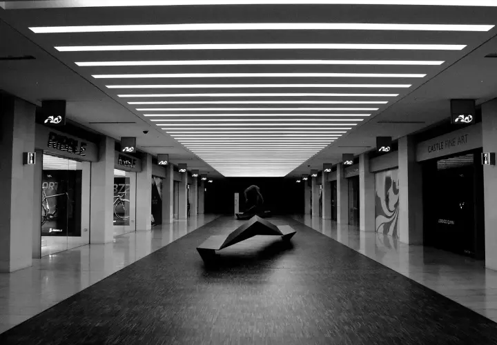 Un couloir de centre commercial, éclairci par des néons, avec une sculpture moderne au centre. Les boutiques sur les côtés sont sombres, créant un contraste saisissant.