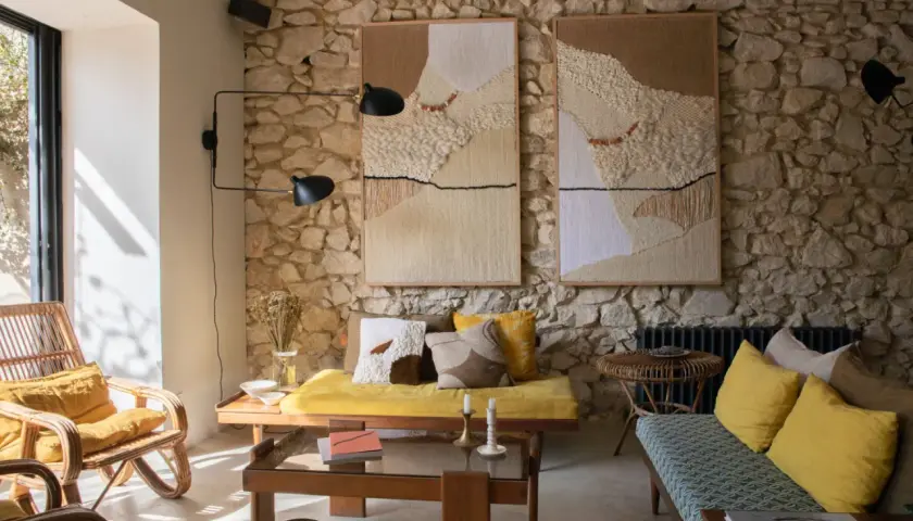 Un salon ancien moderne avec des tapisseries texturées sur un mur de pierre, des meubles en rotin et des coussins jaune vif.