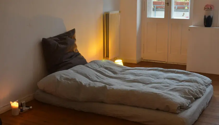 Un matelas au sol sans sommier, prêt pour une nuit paisible. Une bougie allumée éclaire doucement la pièce.