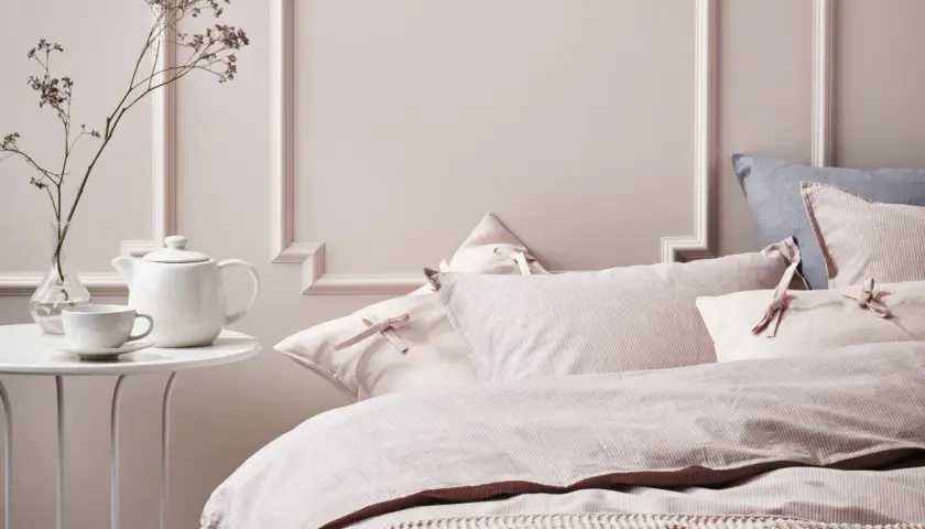 Chambre couleur pastel mur avec un lit douillet, des coussins roses à rayures et une théière blanche sur une table.