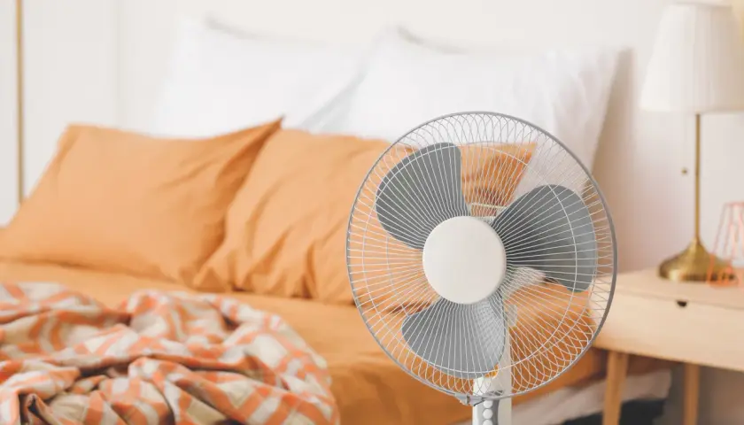 Ventilateur sur pied prêt à rafraîchir une pièce, avec un lit défait en arrière-plan.