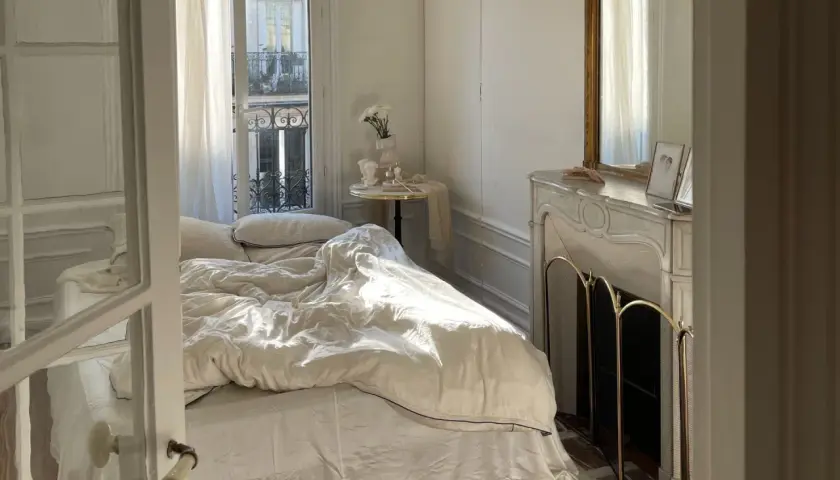 Une petite chambre cocooning baignée de soleil. Le lit défait invite au repos, une cheminée ornée de bibelots et un miroir doré complètent ce tableau paisible.
