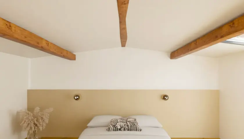 Chambre mansardée aux poutres apparentes, peinte en deux couleurs : blanc et beige. Le lit est fait, avec un coussin décoratif.