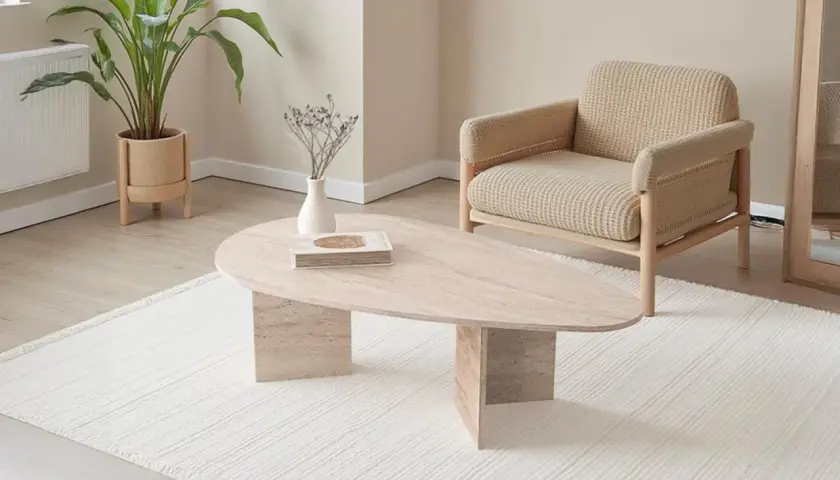 Table basse ovale en travertin, avec une plante verte et un fauteuil en tissu beige.