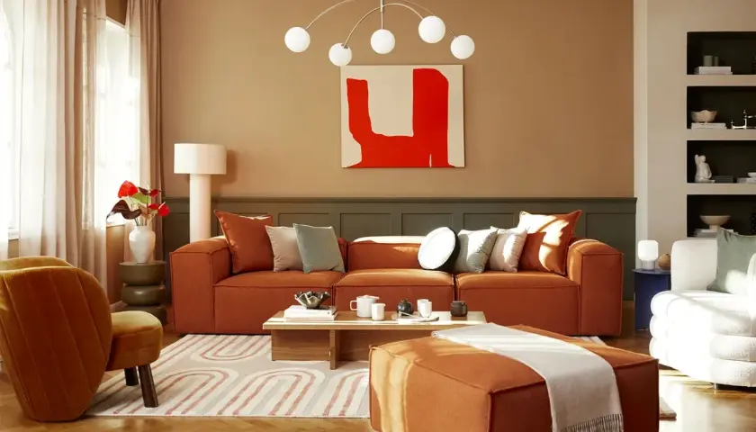 Salon cosy aux tons terreux. Un canapé orange vif, des coussins doux et une peinture abstraite rouge créent une ambiance chaleureuse. La **couleur chaude peinture** domine, invitant à la détente.