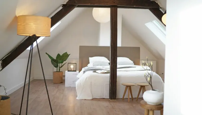 Une chambre minimaliste sous les combles, avec un lit blanc douillet, des poutres apparentes et une plante verte.