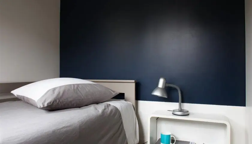 Chambre bleu nuit et taupe : un lit douillet avec un oreiller blanc et gris, une lampe de bureau argentée et une tasse bleue sur une table de chevet blanche.