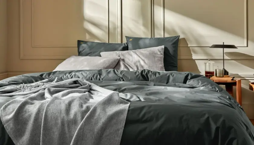 Un lit douillet avec un drap en soie ou satin anthracite, des coussins gris et une couverture douce. La lumière du soleil crée des ombres sur le mur.