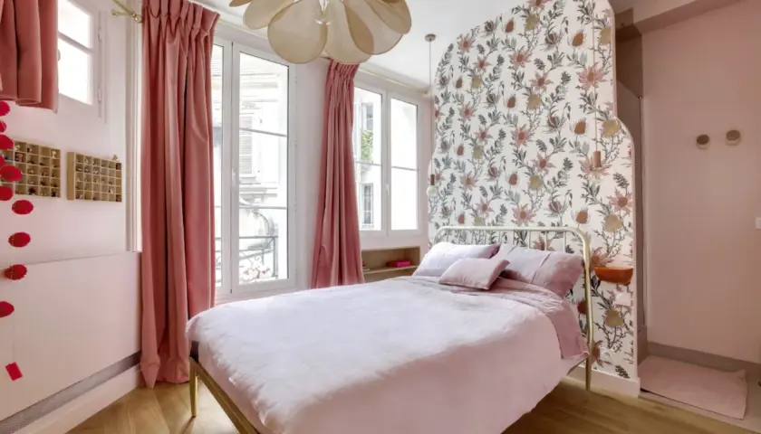 Chambre rose poudré et bois, avec un lit douillet, des rideaux velours, un papier peint floral et un lustre original.