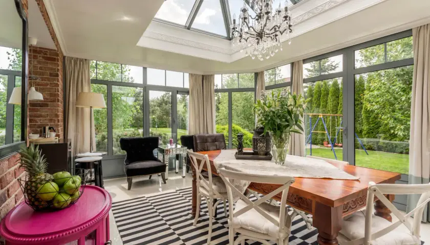 Veranda salon cocooning avec table en bois, lustre en cristal et vue sur jardin verdoyant.