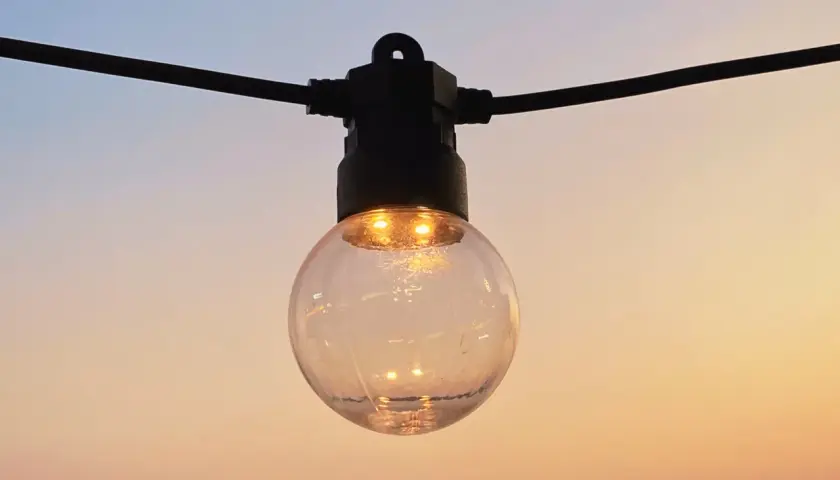 Une ampoule ronde, éclairée, suspendue à un fil noir, se reflète dans le ciel doux du soir. IP 45.